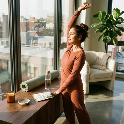 Woman stretching by floor-to-ceiling windows in a modern apartment with a PURETREX D3 K2 supplement pouch on a walnut credenza — Hormonal & Metabolic Health collection